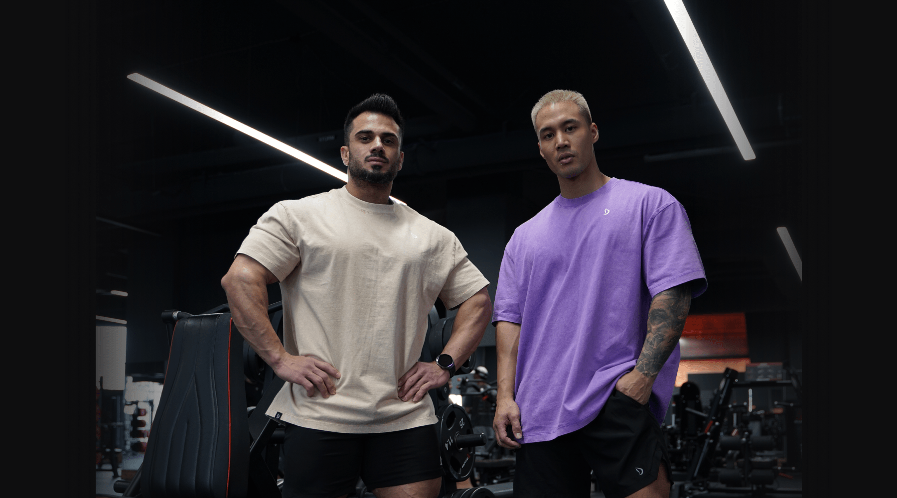 Two lifters in oversized Devoteewear tees, beige and purple, full drape visible, standing in a dark gym